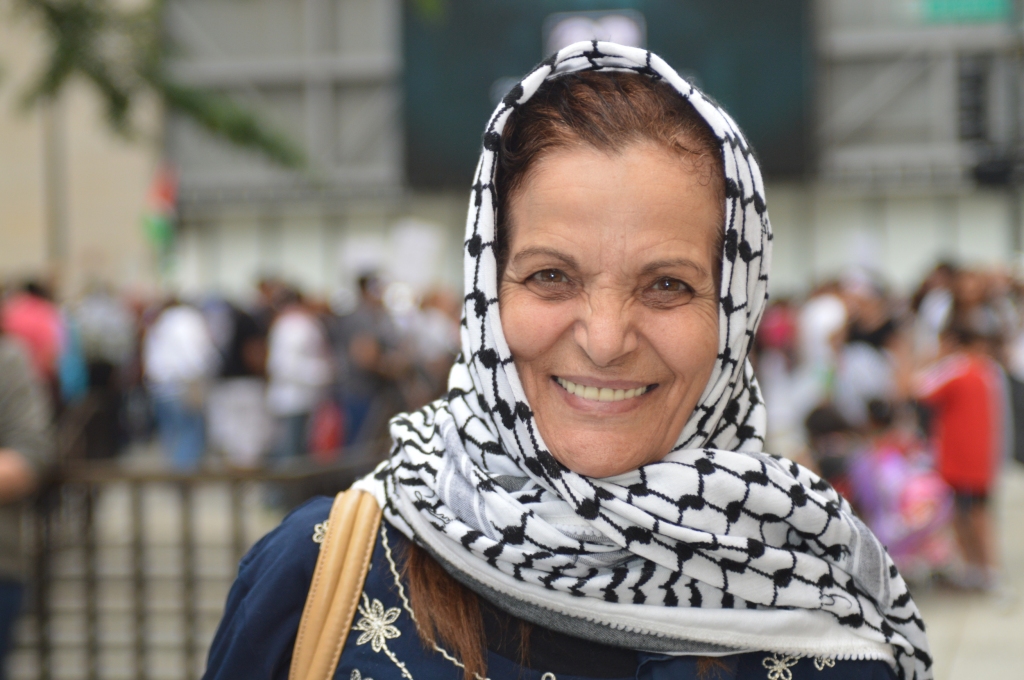 Photo of Rasmea Odeh, a middle aged woman with brown hair mostly concealed under a keffiyeh being worn as a headscarf. She's smiling big enough at the viewer that it crinkles her nose.